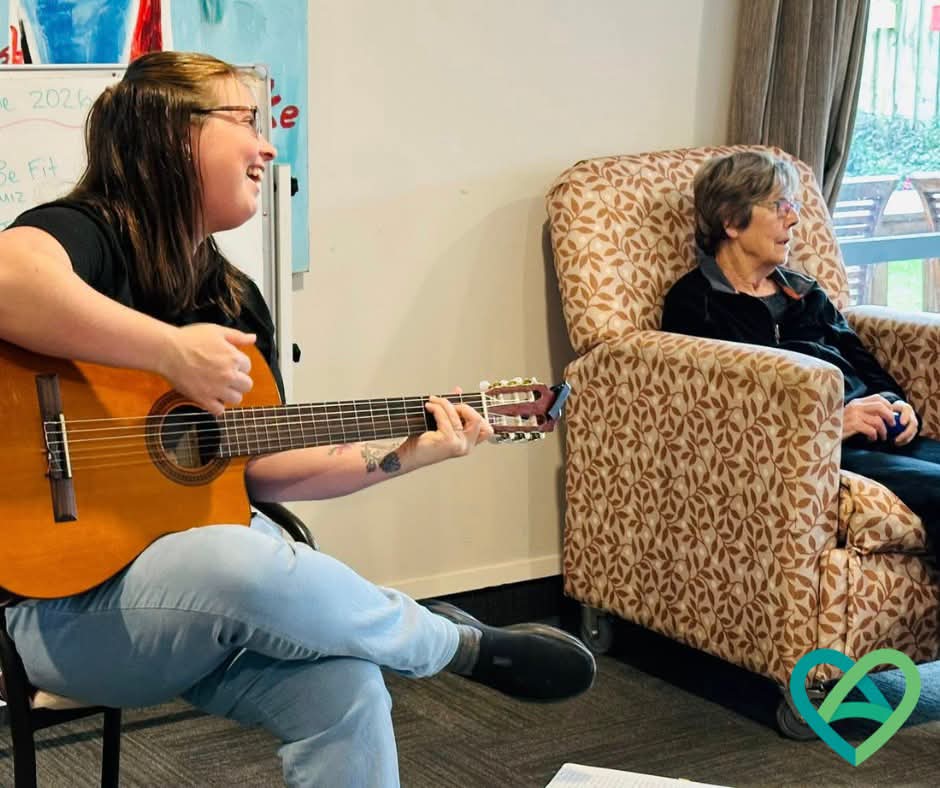 Joanna smiling as she plays guitar beside a resident seated in an armchair